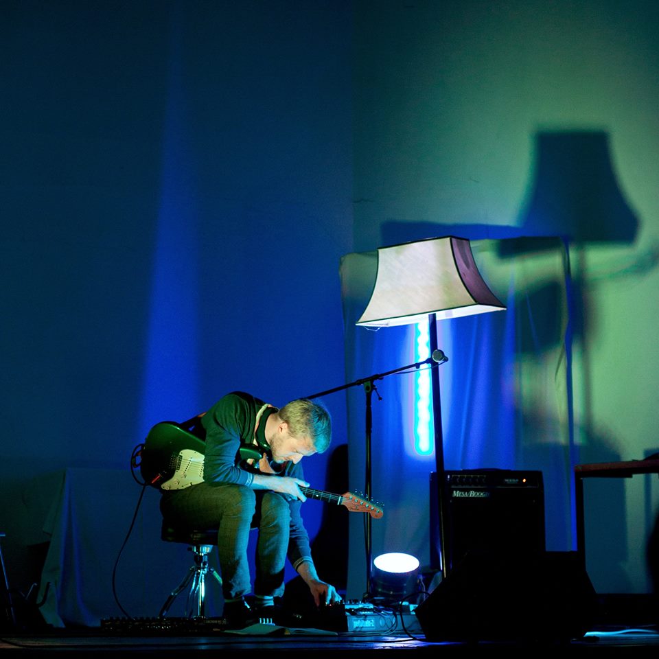 Musician seated on stage playing a guitar under blue and green stage lighting, with a lamp prop beside him, moody atmosphere.